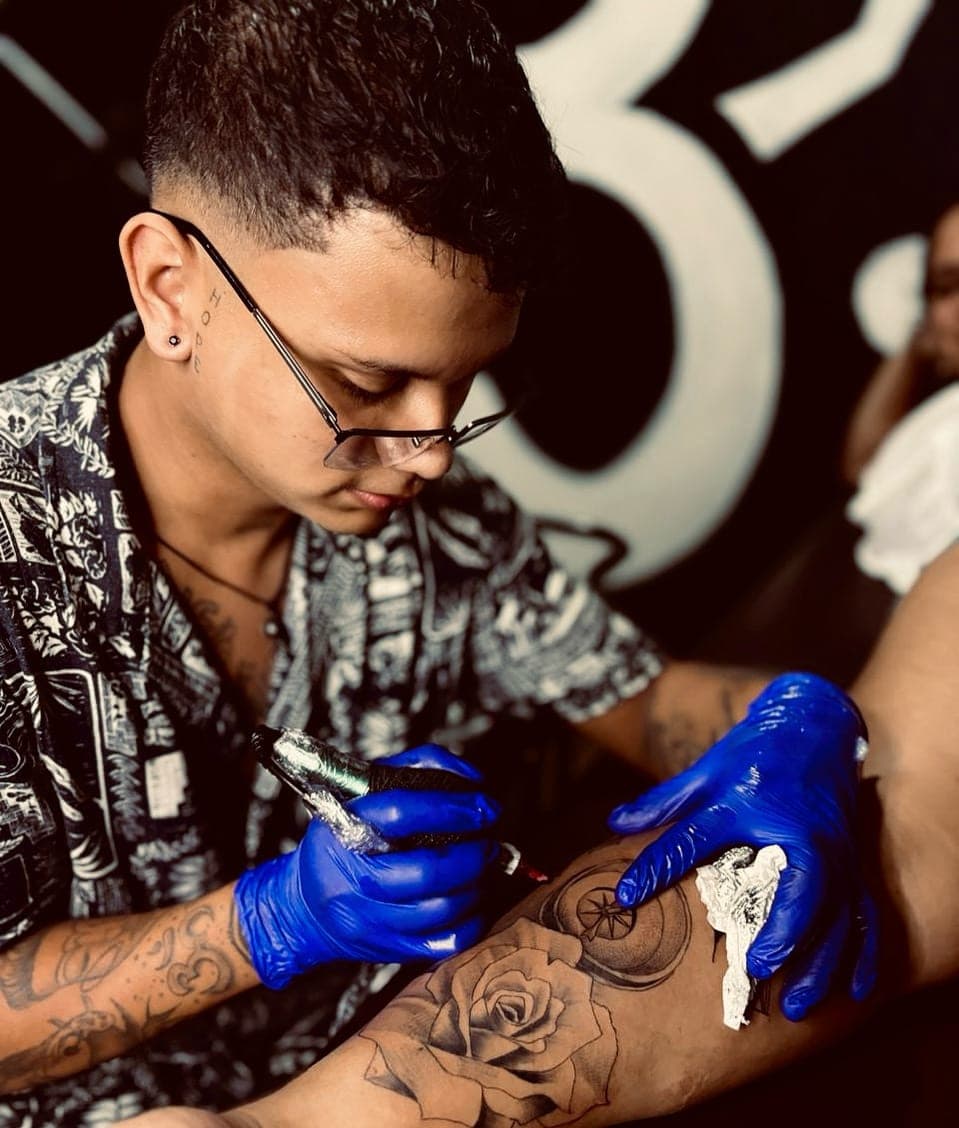 Javier Mena tattooing at the studio, gloved and holding the machine, working a rose on a client's forearm